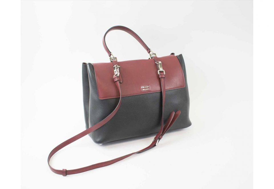PRADA BURGUNDY AND BLACK LEATHER TOTE, with detachable shoulder strap, top  handle, silver tone hardware and bottom feet, front closure with buckles  and twist locks, burgundy leather lining, two main compartments and